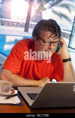 WORKING HOLIDAY KOMMUNIKATIONSTECHNIK URLAUB LAPTOP PROJEKTMANAGEMENT Reife Mann in entspannender Umgebung mit seinem iPhone Smartphone und Laptop-Computer in sonnenbeschienenen Luxus Urlaub Pool Fitness-Raum Situation Stockfoto