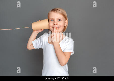 Kleine Mädchen stehen isoliert auf graue Wand Dose halten können Telefon hören, Mund suchen Kamera Lächeln aufgeregt Stockfoto