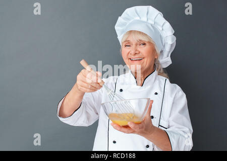 Senior Köchin Studio stehen auf graue Wand rühren, Eier in der Schale kochen isoliert Omelett zum Frühstück suchen Kamera lächelt glücklich close-up Stockfoto