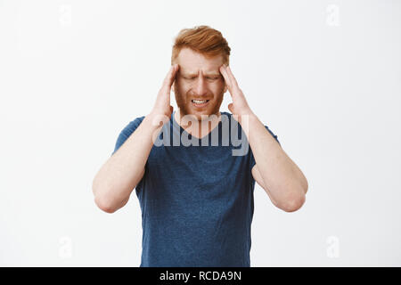 Redhead männliche Unternehmer mit dem roten Haar berühren Tempel runzelte die Stirn und Grimassen von schmerzhaften Kopfschmerzen, Gefühl, Kopfschmerzen oder Migräne, wartet Frau Schmerzmittel zu bringen, stehen über grauer Hintergrund Stockfoto