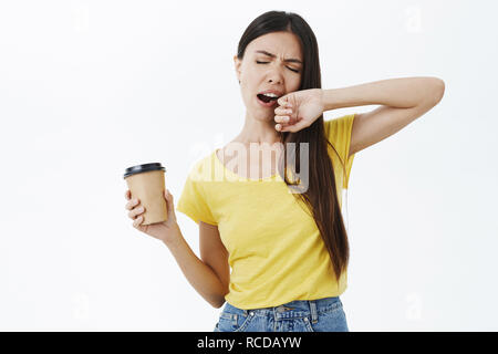 Nur Kaffee kann schlafen kämpfen. Müde und schläfrig Weiblich Mitarbeiter in gelben T-Shirt gähnen mit geschlossenen Augen, öffnete den Mund mit Arm holding Papier Tasse Koffein Aufwachen auf Weg zum Büro Stockfoto