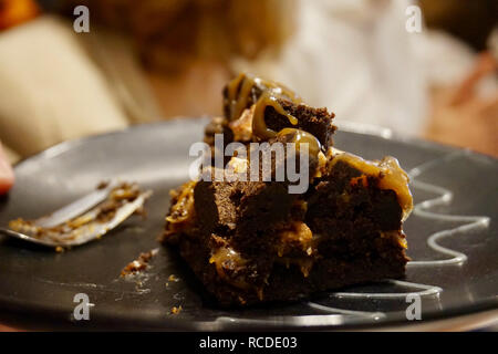 Dichten Schokoladenkuchen mit Karamell, teilweise gegessen und eine schmutzige Gabel im Hintergrund. Auf einer schwarzen Platte, in einer schwach beleuchteten Café. Nahaufnahme. Stockfoto