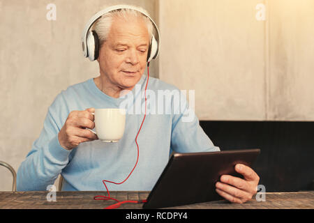 Fesselnde Handlung. Angenehme älterer Mann am Tisch sitzen, Tee trinken und sich einen Film auf die Tablette im Kopfhörer Stockfoto