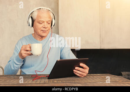 Interessanter Film. Angenehme älterer Mann am Tisch im Speisesaal, einen Film im Tablet in den Kopfhörern beim Tee trinken. Stockfoto