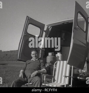 1960s, historisch, oh die einfachen Freuden des Lebens ....ein Mann entspannt sich in einem Klappstuhl auf der Rückseite seines Morris Commercial J4 van auf einem Feld mit seinen Hintertüren weit geöffnet, England, Großbritannien Stockfoto