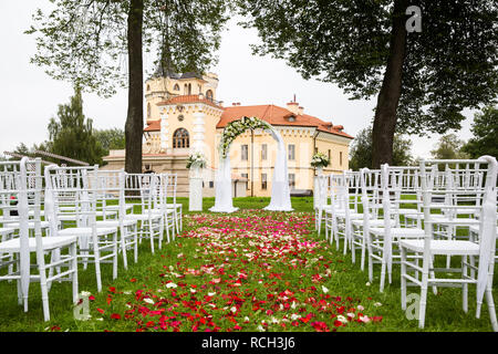 Hochzeit Bogen mit Tuch und Blumen im Freien eingerichtet. Hochzeit auf dem grünen Rasen, bedeckt mit Rosenblättern vor dem Hintergrund des Schlosses Stockfoto