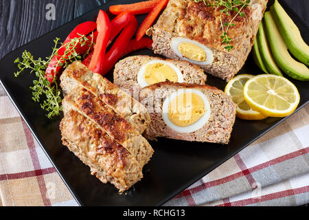 Ansicht von oben der Hackbraten in Scheiben geschnitten serviert mit rotem Pfeffer und Avocado Schichten auf einem schwarzen rechteckigen Platte auf einem Holztisch, Ansicht von oben, close-up Stockfoto