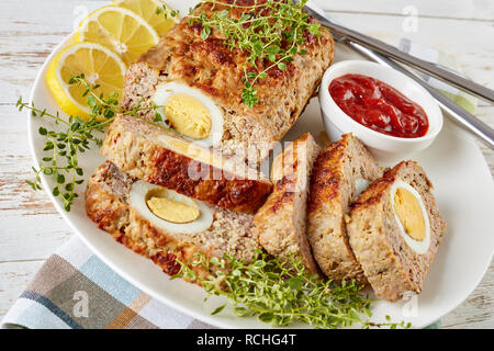 Scheiben Hackbraten gefüllt mit hartgekochten Ei mit Tomatensauce und Zitronenscheiben serviert auf einem weißen Teller mit Messer und Gabel auf einem alten Holztisch, v Stockfoto