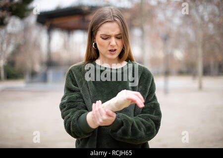 Schöne junge Dame im stilvollen Outfit mit gerunzelter Stirn berühren und Verband auf verletzten Arm beim Stehen im Herbst Park Stockfoto