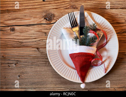 Dekorative Christmas Place Einstellung auf einem rustikalen Tisch mit einem festlichen rote Weihnachtsmütze mit einer Serviette, Dekorationen, Messer und Gabel in einer Ansicht von oben nach unten gefüllt Stockfoto