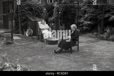 1950s, historisch, eine Frau und ihre Mutter sitzen auf Stühlen in einem Garten, mit der älteren Dame, die ein Buch liest, England, Großbritannien. Stockfoto