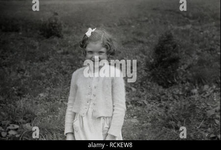 1950s, historisch, ein süßes kleines Mädchen mit Strickjacke und Haarschleife, das draußen auf einem Feld auf dem Land, England, für ein Foto steht. Stockfoto