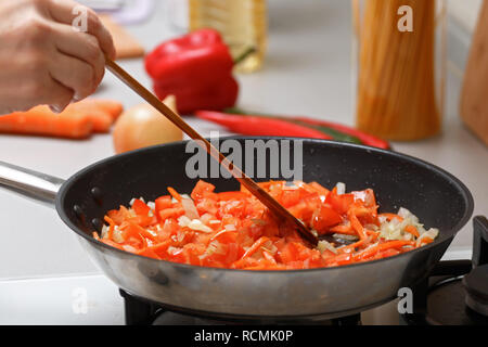 Hausmannskost. Die Frau ist das Rühren der Zwiebeln, Möhren und Tomaten in einer heißen Pfanne mit Pflanzenöl mit einem Holzspachtel entfernen. Close-up. Stockfoto