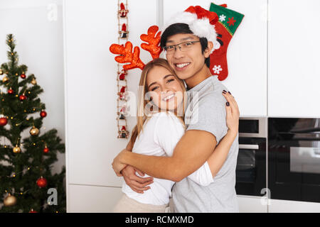 Bild von Paar schöne Mann und Frau tragen Weihnachten Outfit stand in hellen Küche und zusammen umarmen Stockfoto