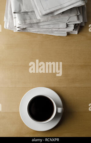 Overhead shot einer Tasse und Untertasse schwarzen Kaffee auf einem hellen Holz Laminat Tisch mit einem Stapel Zeitungen platziert. Stockfoto