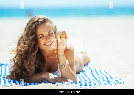 Gerne trendy Frau in weißen Badeanzug am Strand Festlegung auf ein gestreiftes Handtuch und Zuhören, seashell. Perfekte shell - Schöne preiswerte Andenken. Sun Stockfoto