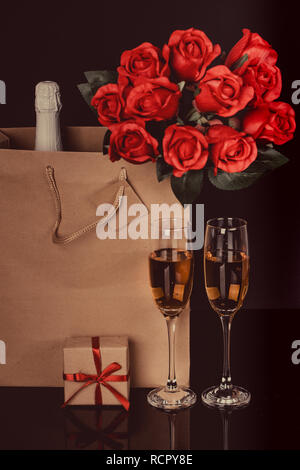 Flasche Champagner und rote Rosen in einer Papiertüte, zwei Weingläser und Geschenkbox auf schwarzem Hintergrund. Liebe, Valentines Tag Konzept Stockfoto