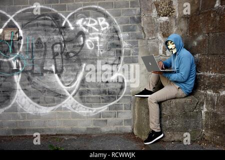 Mann mit einer Maske ist das Surfen auf einem Laptop vor Graffiti Wand bedeckte, symbolische Bild für Hacking Stockfoto
