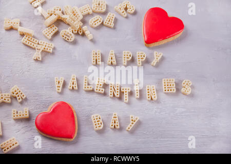 Text Valentines Tag mit Makkaroni in Form von Buchstaben und zwei roten Herzen auf grau Holztisch geschrieben Stockfoto