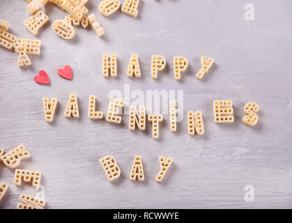 Text Valentines Tag mit Makkaroni in Form von Buchstaben und zwei roten Herzen auf grau Holztisch geschrieben Stockfoto