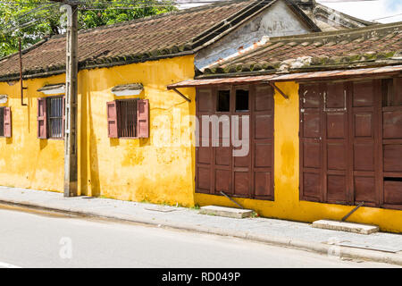 Schrägansicht der hellen gelben Gebäude mit braunen Fensterläden direkt neben der Straße mit Baum im Hintergrund und Strommast in Hoi An, Vietnam Stockfoto