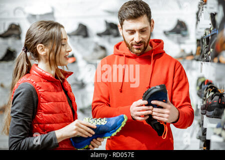 Mam und Frau in Rot Sport Kleidung wählen trail Schuhe für Wanderungen stehen in der Nähe des showacase des modernen Sports Shop Stockfoto