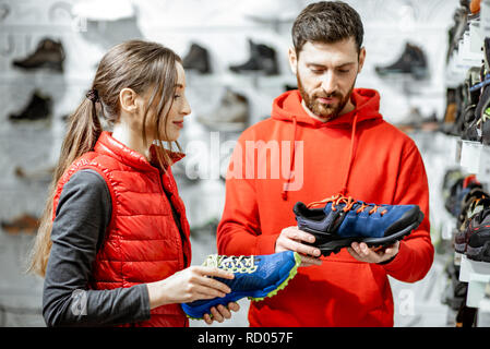 Mam und Frau in Rot Sport Kleidung wählen trail Schuhe für Wanderungen stehen in der Nähe des showacase des modernen Sports Shop Stockfoto