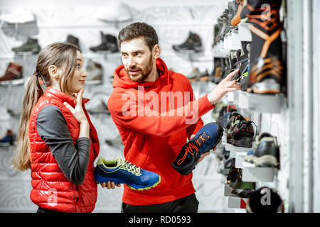 Mam und Frau in Rot Sport Kleidung wählen trail Schuhe für Wanderungen stehen in der Nähe des showacase des modernen Sports Shop Stockfoto