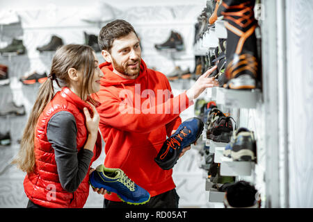 Mam und Frau in Rot Sport Kleidung wählen trail Schuhe für Wanderungen stehen in der Nähe des showacase des modernen Sports Shop Stockfoto