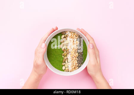 Woman's Hände halten Spinat smoothie Schüssel mit Müsli, Kürbiskerne, Quinoa und Kokos Chips auf rosa Hintergrund. Ansicht von oben minimal gehalten. Stockfoto