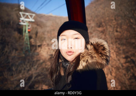 Eine junge chinesische Frau in einem Skilift Gondel sitzen Tragen schwerer Winterkleidung bläst einen Kuss. Stockfoto