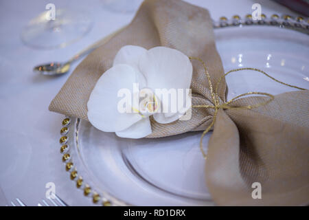 Fine Dining table Setting mit transparenten Platten, beigefarbene Bettwäsche Serviette mit natürlichen Orchideen und goldenen Dekorationen für Gäste bei einer Hochzeit bereit Stockfoto