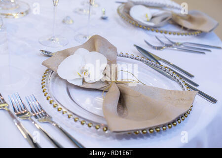 Fine Dining table Setting mit transparenten Platten, beigefarbene Bettwäsche Serviette mit natürlichen Orchideen und goldenen Dekorationen und glänzendes Besteck Stockfoto