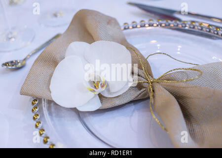 Fine Dining table Setting mit transparenten Platten, beigefarbene Bettwäsche Serviette mit natürlichen Orchideen und goldenen Dekorationen für Gäste bei einer Hochzeit bereit Stockfoto