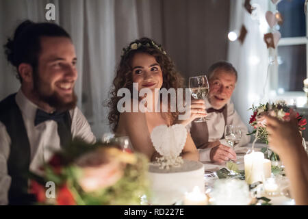 Ein junges Paar, das an einem Tisch auf einer Hochzeit sitzen, im Gespräch mit Gästen. Stockfoto