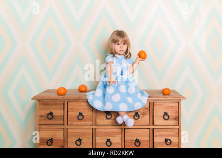 Kleines Mädchen mit überkreuzten Beinen auf Kommode holding Tangerine in der Hand und unten mit Zeigefinger auf Raute Hintergrund. Kind in blau gepunkteten Kleid und weißen Socken Stockfoto