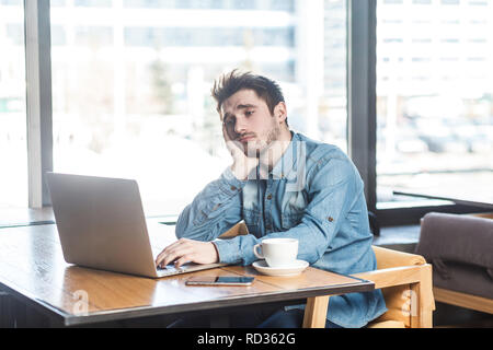 Wissen Sie nicht! Portrait von Traurigkeit müde bärtige junge Freiberufler in Blue Jeans shirt sind allein im Cafe sitzen und auf der Suche nach einer Arbeit auf dem Laptop sind in Stockfoto
