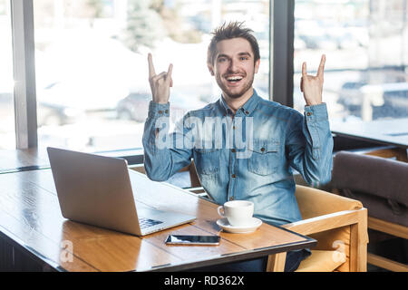 Portrait von Stattlichen erfolgreich positive bärtige junge Freiberufler in Blue Jeans shirt Sitzen im Cafe und Arbeiten am Laptop mit toothy Lächeln und Stockfoto