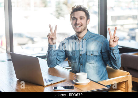 Portrait von Stattlichen erfolgreich positive bärtige junge Freiberufler in Blue Jeans shirt Sitzen im Cafe und Arbeiten am Laptop mit toothy Lächeln und Stockfoto