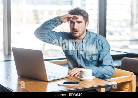 Portrait von Stattlichen bärtige junge Freiberufler in Blue Jeans shirt fragte sich, Sitzen im Cafe und Arbeiten am Laptop, die Hand in der Nähe der Stirn mit Stockfoto