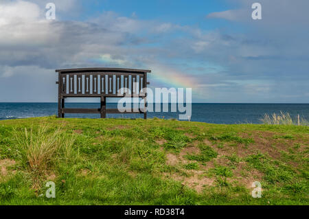 Eine Bank mit einem Regenbogen über der Nordsee Küste, im Benthall, Northumberland, England, UK gesehen Stockfoto