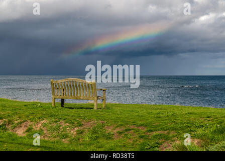 Eine Bank mit einem Regenbogen über der Nordsee Küste, im Benthall, Northumberland, England, UK gesehen Stockfoto