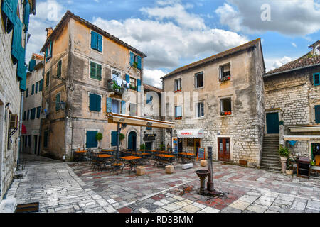 Einen kleinen Platz in der Altstadt von Kotor, Montenegro, mit einer Wasserpumpe in der Mitte von einem Cafe, Markt und Apartments mit bunten Fenstern umgeben. Stockfoto