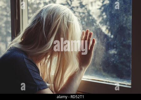 Blonde Frau lehnte sich gegen Fenster, Blick, traurig, deprimiert und einsam mit unbrushed lange Haare, nicht sichtbare Antlitz Stockfoto