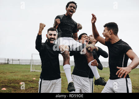 Fußballspieler schreien vor Freude und pumpen nach einem Sieg ihre Fäuste. Spieler, die auf dem Spielfeld unterwegs sind und einen Teamkollegen auf den Schultern tragen, der einen Sieg feiert. Stockfoto