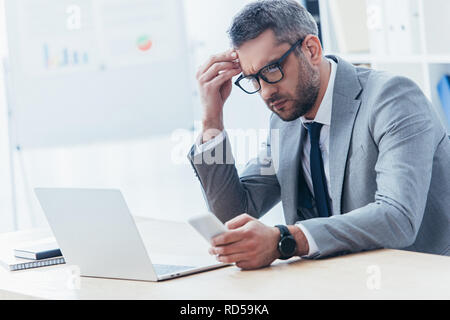 Konzentrierte Geschäftsmann in Brillen mit Smartphone und Arbeiten mit Laptop im Büro Stockfoto
