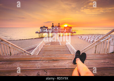 Junge Mann sitzt auf hölzernen Pier genießen Sie einen wunderschönen Sonnenaufgang am berühmten Seebrucke Sellin in Ostseebad Sellin, Insel Rügen, Ostsee, Deutschland Stockfoto