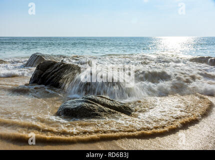 Eine kleine Welle von einem ruhigen Meer stürzt über grosse Felsen an einem Sandstrand. Stockfoto