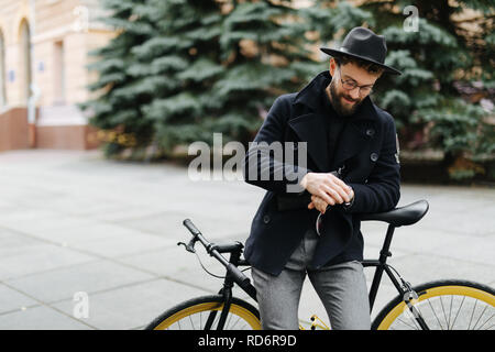 Gut aussehender bärtiger Mann, der einen Bruch sitzt auf einem Fahrrad auf die Uhr schauen. Stockfoto