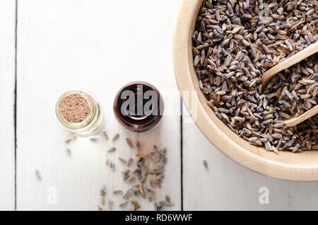 Overhead shot von eine hölzerne Schüssel mit getrocknetem Lavendel und zwei Durchstechflaschen aus Glas mit Öl gefüllt. Weitere Knospen auf weißem Holz beplankt Tisch verstreut. Stockfoto